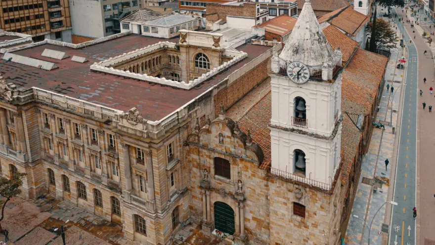  Iglesia San Francisco colonial en primer plano de fachada con blanca torre del reloj y techo, al lado se observa la cicloruta de la carrera séptima con algunas personas caminando.