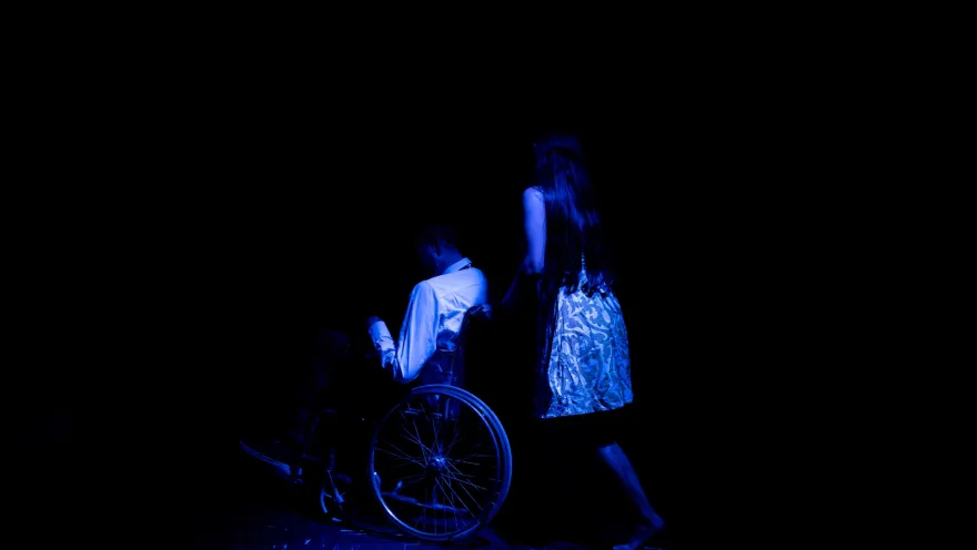  Barraca Teatro con actor en silla de ruedas en escena con luz azul. Una mujer lo mueve en la silla.