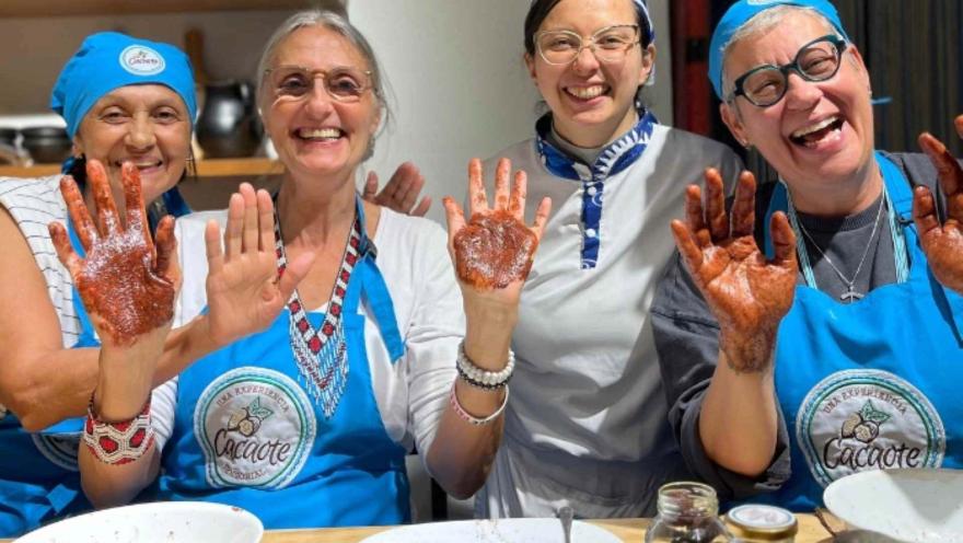 Cuatro mujeres, tres adultas y una joven con delantales y gorros azules sonríen y muestran sus manos untadas de cacao.