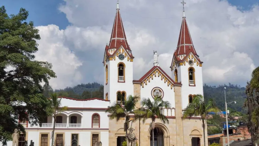 Iglesia Parroquial San Miguel Arcángel - Gachetá fachada de frente en parque rodeada de árboles  y obreros que caminan al frente. Al fondo se observan montañas y un cielo azul con nubes blancas.