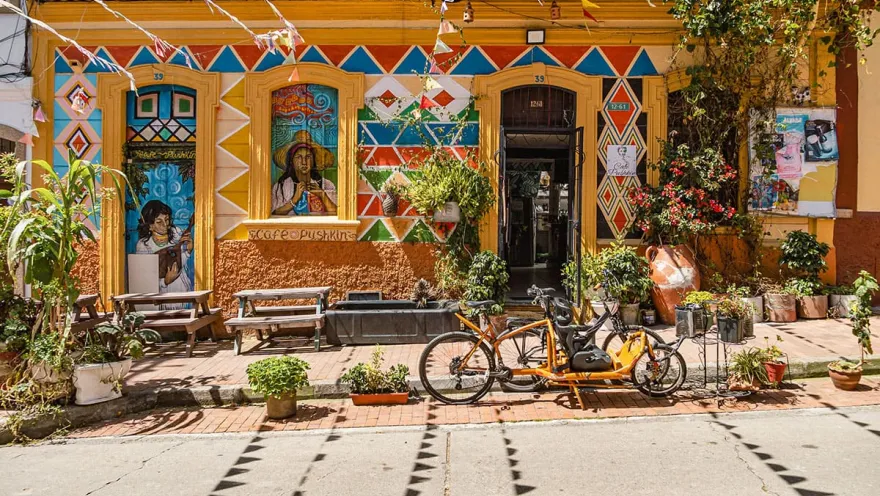 La Candelaria – Facade of a one-story building with colorful graffiti in yellow, blue, and red depicting farmers. The door is open, with bicycles parked in front, surrounded by flowerpots and plants.