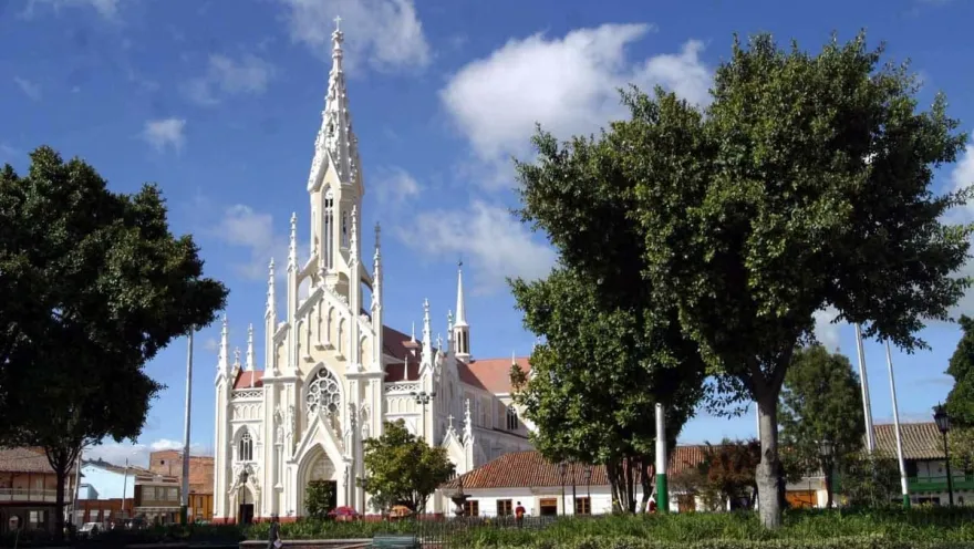 Parroquia Basílica Menor del Santo Cristo - Ubaté neogótica en parque rodeada de árboles y cielo azul con una nubecita blanca. Se en dos personas caminando a lo lejos.