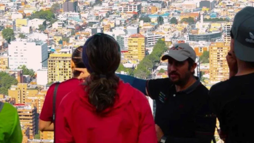 Grupo de personas de espalda observan la ciudad desde un mirador, mientras que un guia hombre parece explicar.