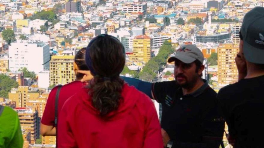 Grupo de personas de espalda observan la ciudad desde un mirador, mientras que un guia hombre parece explicar.