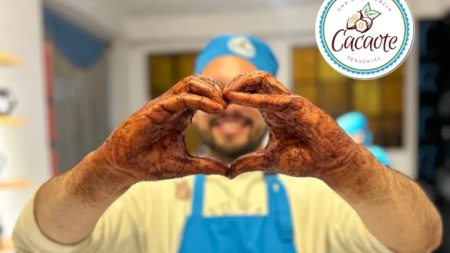 Un hombre joven con delantal y gorro azul muestra sus manos untadas de cacao y haciendo una forma de corazón.