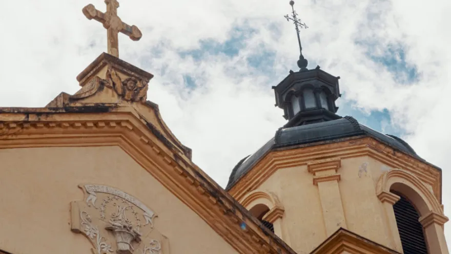 Iglesia de La Candelaria, parte superior de una de las torres de color entre amarillo y negro terminada en cúpula con cruz metálica negra mirando al cielo azul con nubes blancas.