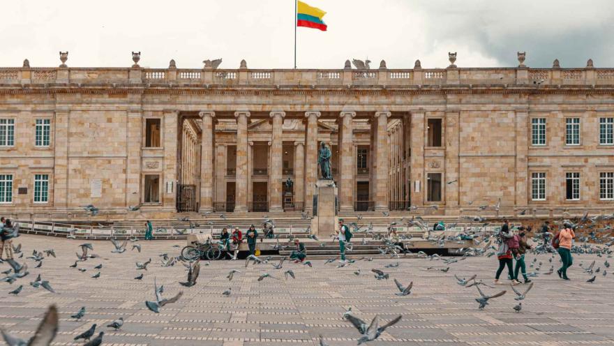 Plaza de Bolívar vista norte de a sur con vuelo de palomas, personas y una estatua de Simón Bolívar, y al fondo el Capitolio nacional donde ondea una bandera de Colombia.