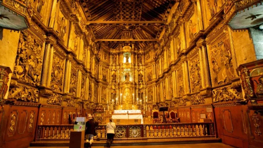  Iglesia San Francisco colonial con su interior, techos y paredes en retablos de madera dorada. Dos mujeres de rodillas oran al frente del altar blanco iluminado con varios velones. Al frente se observan figuras de varios santos iluminados con luz artificial.  
