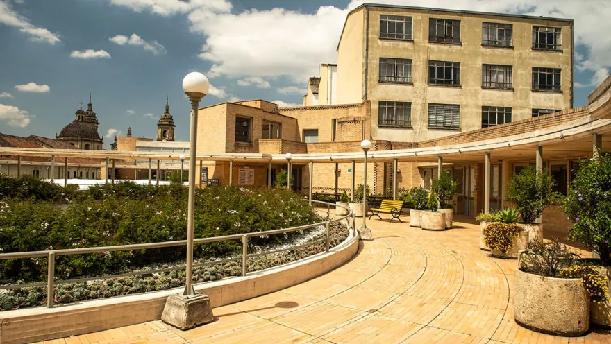 La Candelaria – Gabriel García Márquez Cultural Center, circular brick corridor with flowerpots, chairs, and light posts. In the background, an old building and the domes of the Primatial Cathedral stand out.