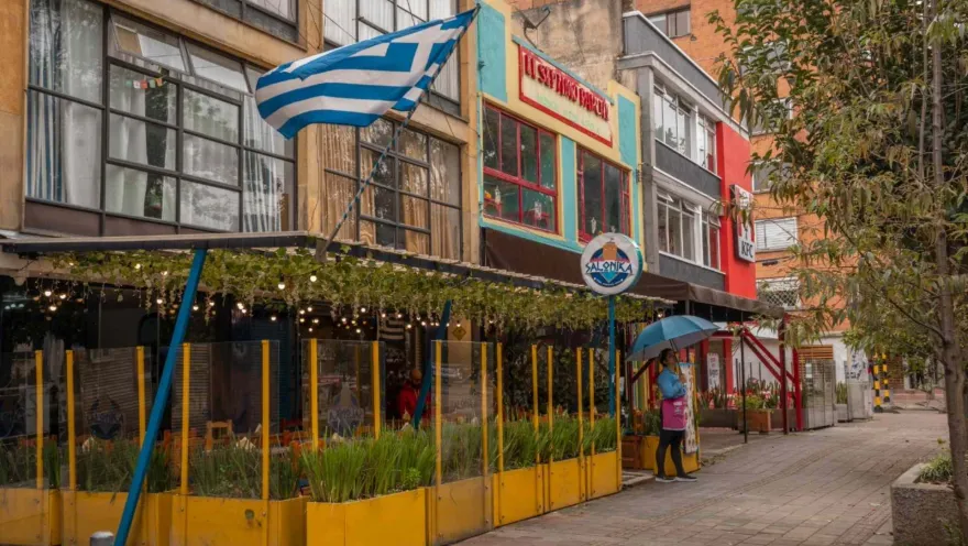 Park Way fachadas laterales del restaurante griego Salónica en la parte baja de un edificio. Hay una bandera de Grecia y una mesera con una sombrilla a la entrada.