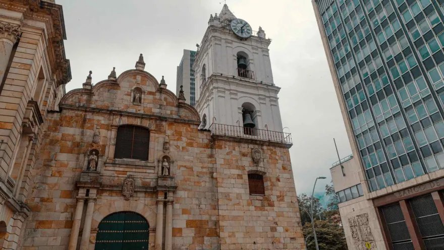  Iglesia San Francisco colonial su fachada vista de frente desde la puerta de madera verde y cerrada, hasta la torre blanca del campanario y reloj. Al lado izquierdo se ve la fachada del edificio del Banco de la República.