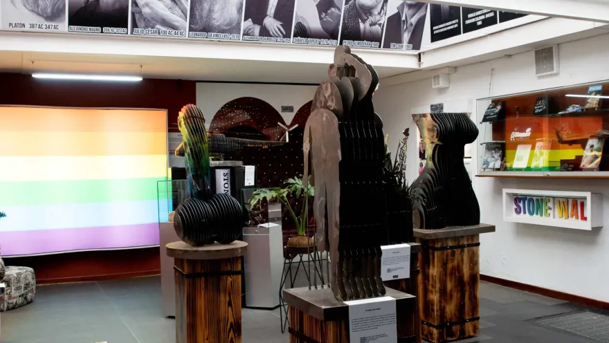 Museo Lgbti al interior salón de exhibición de obras en cobre, una con forma de falo gigante y otra un cuerpo de mujer. Hay fotografías de personalidades gay históricas y una pantalla con los colores de la bandera arcoíris lgbti.  