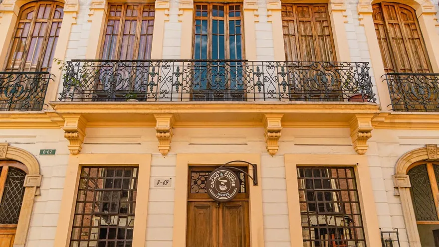La Candelaria – Republican-style building façade with five wooden windows on the second floor, a wide balcony with metal railings, and on the first floor, a large door and two windows. A small round sign reads “Cacao & Coffee House.”
