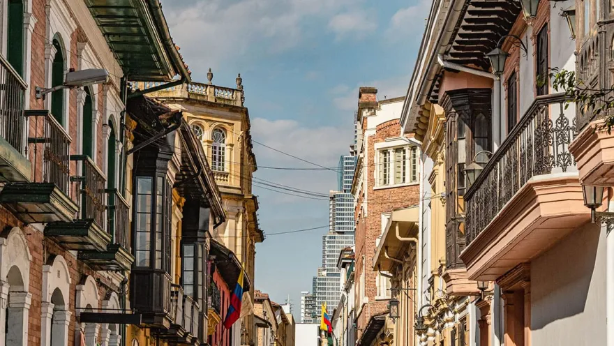 La Candelaria – View of a colonial street with houses featuring balconies and a pair of Colombian flags. In the background, the modern Bacatá building can be seen.