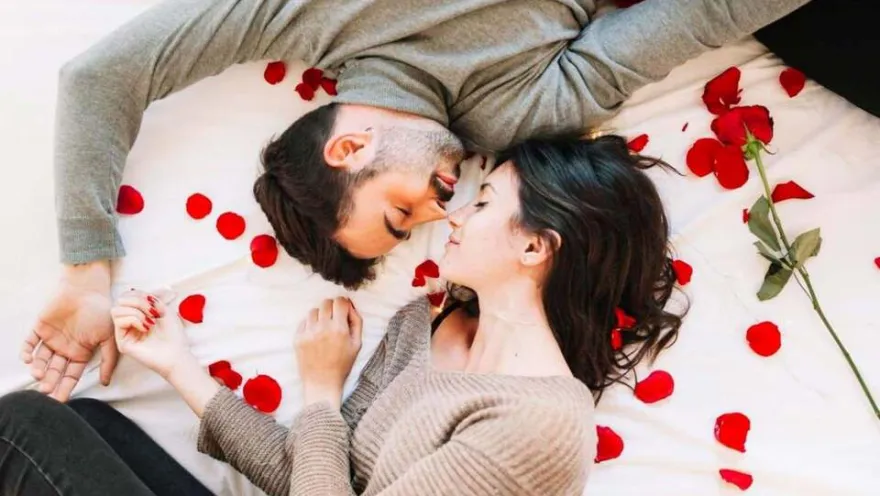 Pareja de hombre y mujer jóvenes recostados sobre una cama decorada con rosas rojas. 