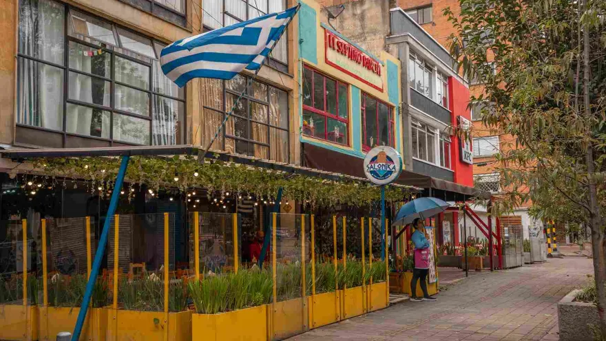 This image shows a vibrant and colorful street on Park Way. In the foreground, the facade of a Greek restaurant called 'Salonika' stands out with its cheerful colors. A metal and glass awning with hanging lights covers the terrace, creating a welcoming atmosphere. The Greek flag waving above the restaurant and the yellow and blue details evoke Mediterranean culture.