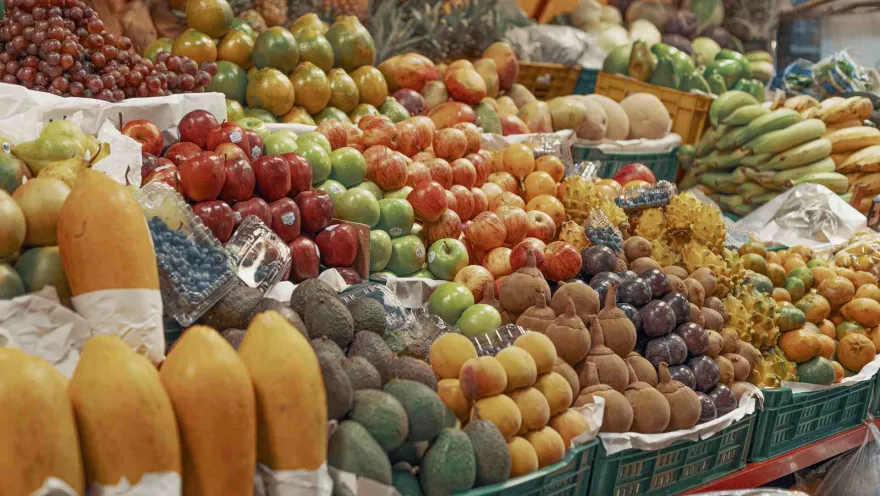 Esta imagen muestra un puesto de frutas muy bien surtido en un mercado. Se pueden ver grandes cantidades de papayas, aguacates, arándanos, uvas rojas y verdes, manzanas (rojas y verdes), así como una variedad de frutas exóticas como kiwanos. Las frutas están organizadas en montones y cestas, creando una exhibición colorida y abundante.