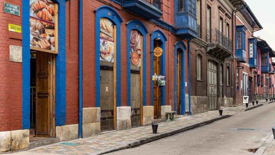 Esta fotografía capta la esencia de La Candelaria, el centro histórico de Bogotá. La imagen muestra una calle empinada bordeada por edificios de colores vibrantes y arquitectura colonial. El edificio principal, de ladrillo rojo con marcos y balcones de un azul intenso, alberga un negocio local con vitrinas que exhiben pan y pasteles