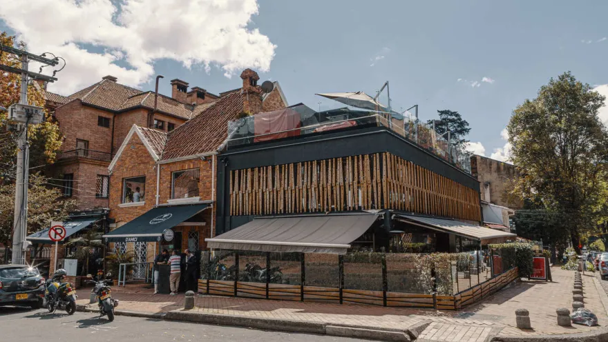 Esta imagen muestra una esquina vibrante de la Zona G en Bogotá. En el centro de la toma, un edificio de dos pisos destaca por su moderna fachada negra con detalles en madera, lo que le da un toque sofisticado y orgánico. El cielo azul con nubes dispersas ilumina el paisaje, creando una atmósfera acogedora.