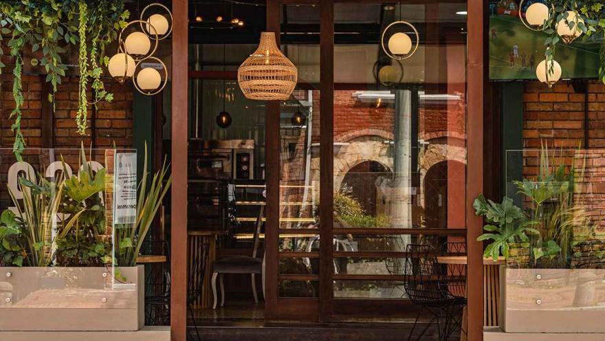 Esta imagen muestra la fachada de un restaurante acogedor la Zona G de Bogotá. La entrada, con sus grandes ventanales de madera, invita a mirar el interior, donde se ven luces cálidas y una decoración moderna. A los lados, plantas verdes en macetas y luces colgantes en forma de esferas y canastas tejidas crean un ambiente sofisticado y natural. El diseño integra elementos rústicos con toques contemporáneos, lo que lo convierte en un lugar ideal para disfrutar de una experiencia gastronómica