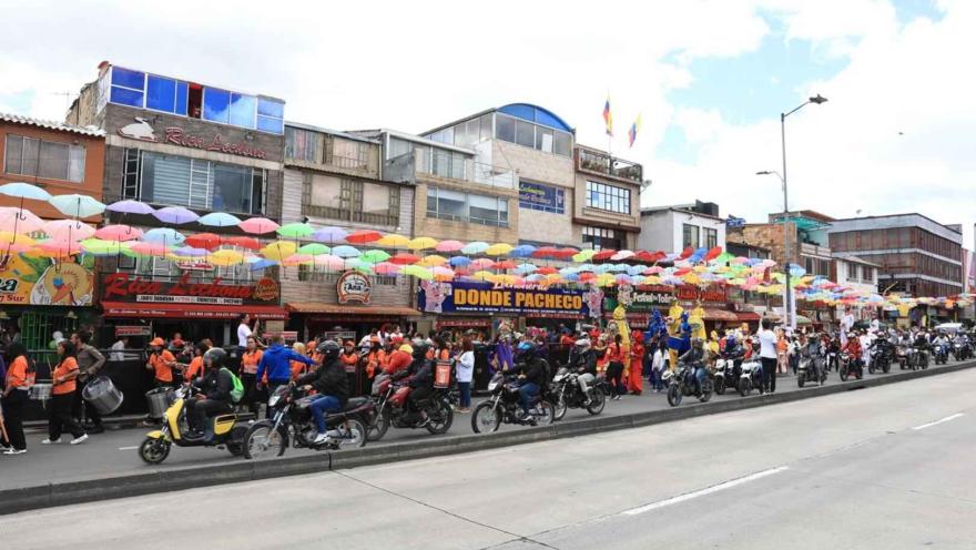 Esta imagen vibrante muestra una calle animada en un sector comercial, posiblemente durante un festival o evento. Una multitud de personas, algunas en motocicleta y otras a pie, se congregan bajo un cielo de paraguas de colores que cuelgan sobre la calle. Los edificios de fachadas variadas, algunos con letreros de negocios locales, crean un fondo dinámico. La presencia de motocicletas y la gente en movimiento sugieren una celebración o un día de actividad comercial intensa, con un ambiente festivo 