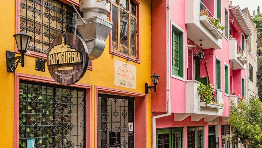 Esta imagen de una calle de La Macarena captura la vibrante mezcla de colores que caracteriza al barrio. En primer plano, se ve una fachada en tonos de naranja, amarillo y rojo, seguida de un edificio en tonos rosados con balcones blancos y ventanas verdes. La diversidad arquitectónica es evidente, con una mezcla de estilos y alturas que le dan al barrio su carácter ecléctico. Las calles empinadas y el follaje de los árboles en el borde de la calle son elementos típicos de este barrio bohemio y artístico.