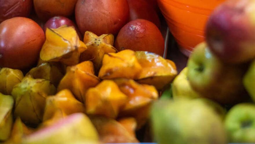 Un primer plano enfocado en una colorida variedad de frutas exóticas y frescas, dispuestas en un puesto de mercado. En el centro, se destacan varias carambolas (frutas estrella) de color amarillo brillante y frutas rojizas. A la derecha, un recipiente naranja contiene más frutas redondas. El fondo y el primer plano están suavemente desenfocados, mostrando una profusión de otras frutas verdes y amarillas, lo que sugiere una abundancia de productos.