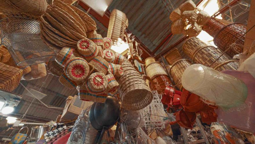 La fotografía ofrece una perspectiva de ángulo bajo, mostrando un techo de mercado desde el cual cuelgan innumerables cestas y objetos de mimbre de diversas formas y tamaños. También se aprecian algunas lámparas, utensilios de cocina metálicos y objetos envueltos en plástico, creando una composición densa y visualmente interesante de productos artesanales y domésticos.