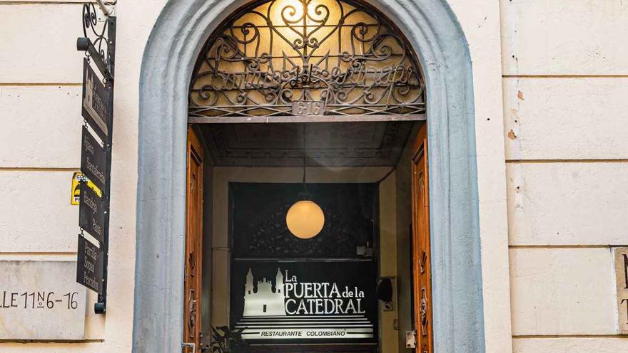Esta imagen de cerca muestra una entrada icónica en el centro de Bogotá. Se trata de 'La Puerta de la Catedral', un restaurante ubicado en el corazón de La Candelaria. La lámpara colgante en el interior y el letrero discreto invitan a los transeúntes a descubrir un lugar lleno de historia y sabor, justo en las calles del centro histórico.