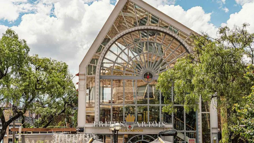 This photograph showcases the imposing architecture of a building in Zona G, which houses a restaurant called 'Frites Artois'. The structure is characterized by its large, arched glass facade with metal details that lend it an industrial and modern feel. The building is surrounded by leafy trees, and in the foreground, the umbrellas of the restaurant terraces invite diners to enjoy the fresh air.