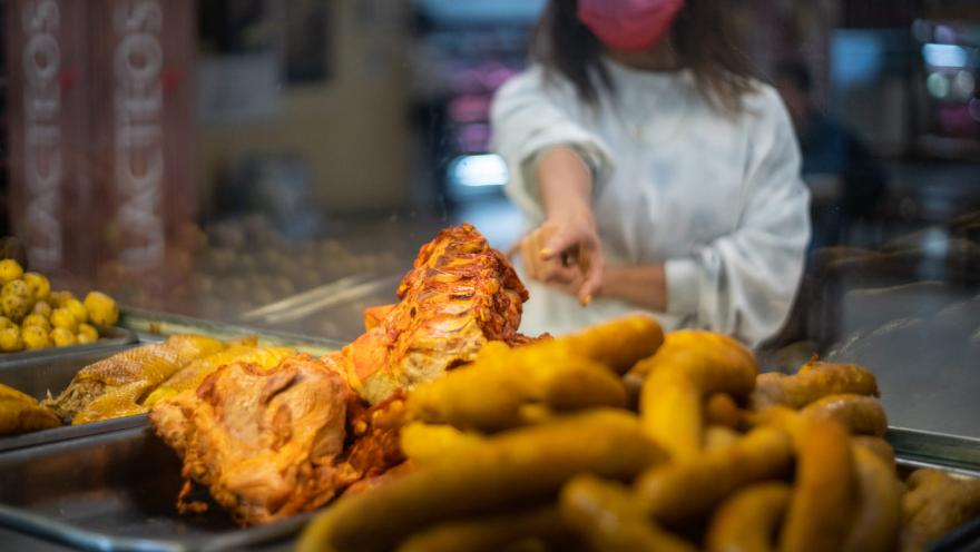 La imagen muestra una vitrina de comida de una Fritanga tradicional. En primer plano, se ven trozos de carne asada o frita, con huesos visibles, y una gran cantidad papas criollas, gallina y chunchullo. Detrás del cristal, una mujer con una mascarilla rosa y una sudadera blanca extiende la mano hacia la comida, sugiriendo que está a punto de servir o tomar un plato.