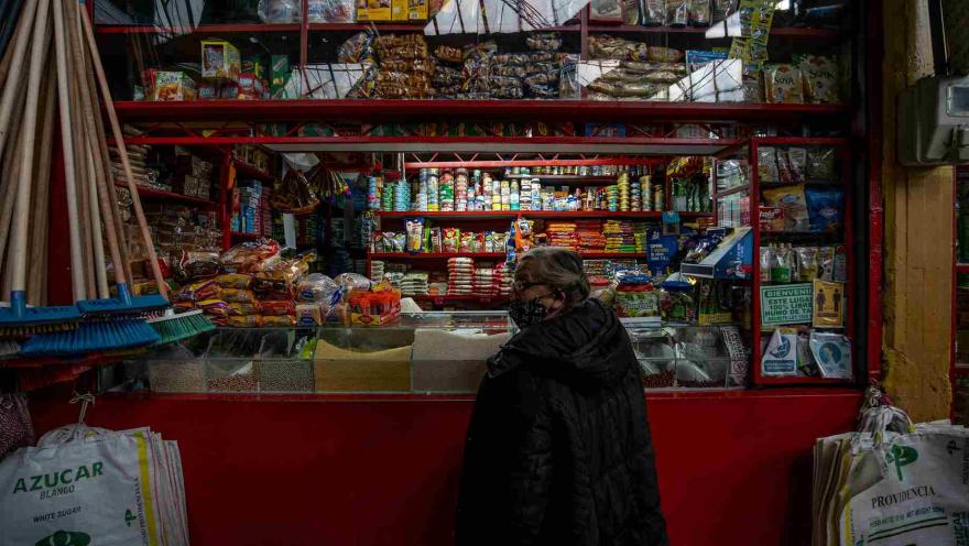  Esta imagen muestra el interior de una pequeña tienda. Una persona con abrigo oscuro está de espaldas, mirando los productos. El puesto está repleto de estantes con una variedad de artículos, incluyendo latas, paquetes de alimentos, escobas y bolsas grandes de azúcar o granos en la parte inferior. La iluminación superior se refleja en una ventana, revelando más productos apilados.