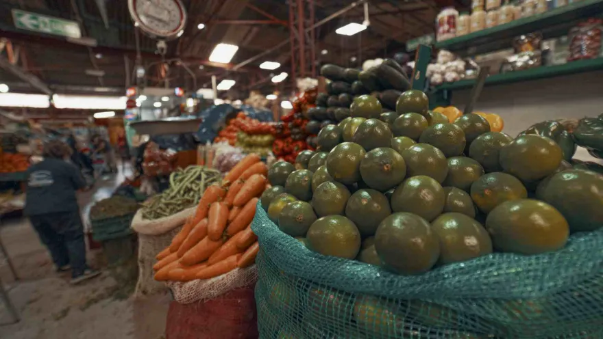 En esta imagen, un primer plano enfocado en grandes sacos de frutas cítricas verdes domina el lado derecho. Detrás y a la izquierda, se ven sacos de zanahorias, calabacines y otros vegetales apilados. Al fondo, se distingue el ambiente bullicioso de un mercado, con una persona caminando y estantes llenos de productos. Una báscula circular se ve desenfocada en la parte superior izquierda.