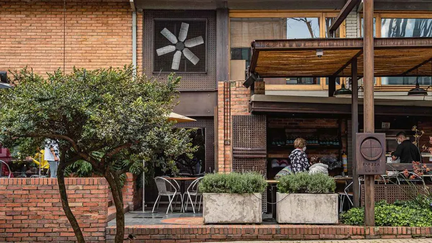 The building, with its brick walls and wood details, features a large window with a ceiling fan that adds a distinctive touch. An outdoor terrace, bordered by concrete planters filled with greenery, invites diners to enjoy the fresh air. The sidewalk bricks and outdoor tables contribute to the relaxed and modern atmosphere.