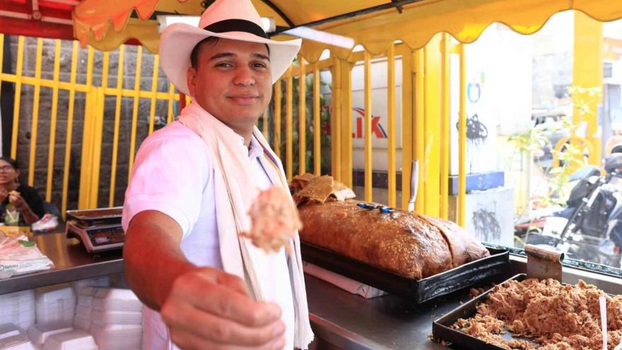 Esta imagen de cerca muestra a un hombre sonriente vestido con un sombrero blanco y una camisa blanca, el atuendo tradicional del tolimense. Está ofreciendo un trozo de lechona, la cual se ve en grandes porciones en bandejas de metal detrás de él. El ambiente es de un puesto de comida al aire libre, con el toldo amarillo y la barra de acero. Su expresión amigable y el gesto de ofrecer la comida transmiten la calidez de la hospitalidad colombiana y la riqueza de su gastronomía.
