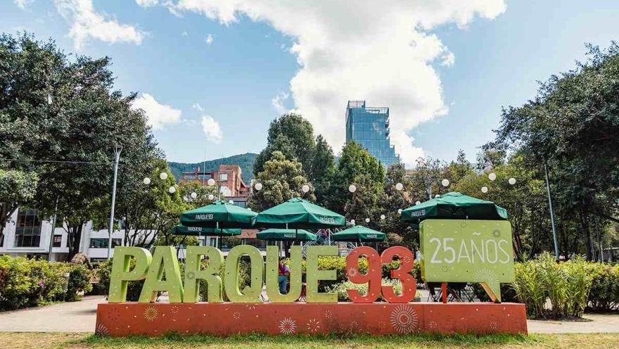 La imagen muestra una toma frontal del letrero icónico del Parque de la 93. La estructura, que celebra los "25 AÑOS" del parque, es una escultura de color naranja con el texto "PARQUE 93" en letras grandes de color verde. Detrás del letrero, se pueden ver las sombrillas de los cafés y restaurantes que rodean el parque, así como los altos árboles y un rascacielos en el fondo. El cielo azul y el clima soleado hacen que el ambiente sea vibrante y lleno de vida.