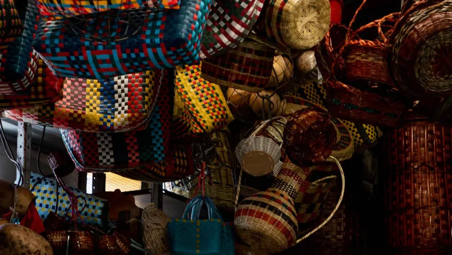 Esta imagen de cerca muestra una colorida colección de cestas y objetos artesanales hechos de mimbre, colgados en un mercado o tienda de artesanías. Los objetos, de diferentes formas y tamaños, están tejidos con patrones geométricos y colores vibrantes, como el rojo, azul, amarillo y verde. La diversidad de los diseños y la habilidad artesanal son evidentes en cada pieza. La imagen evoca la riqueza cultural y la tradición de los artesanos locales.