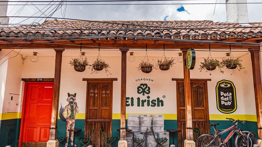 Entrada del pub “El Irish” en Usaquén, decorada con un mural artístico de un zorro elegante y un diseño que mezcla tradición e identidad irlandesa. Su fachada blanca con tejas de barro, mesas en la parte exterior y bicicletas estacionadas transmiten un ambiente bohemio y relajado.
