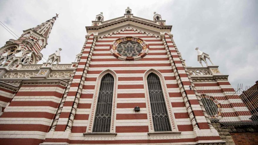 Santuario Nuestra Señora del Carmen en primerísimo primer plano de fachada gótica vista desde abajo sobresaliendo las franjas horizontales rojas y blancas, los adornos del techo y la aguja de la torre principal. El cielo es gris.