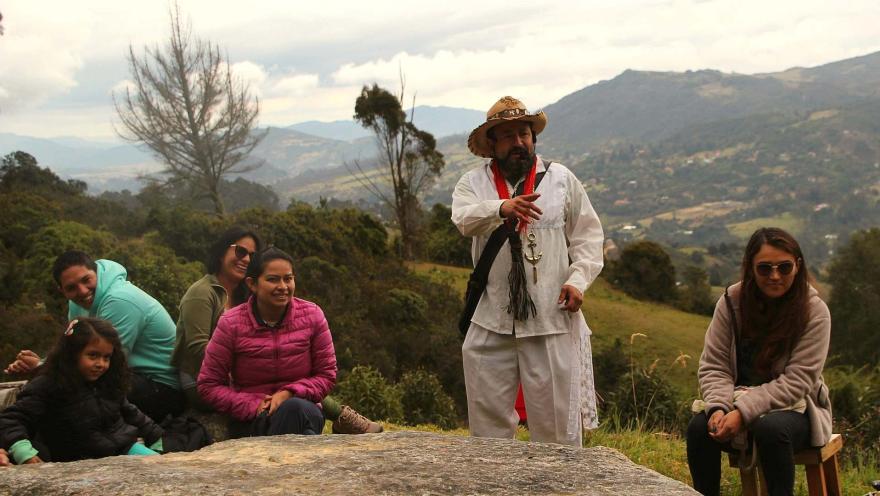 Grupo de personas, con ellos una niña, en el campo, rodeados por montañas. Un hombre vestido como campesino está hablando. 