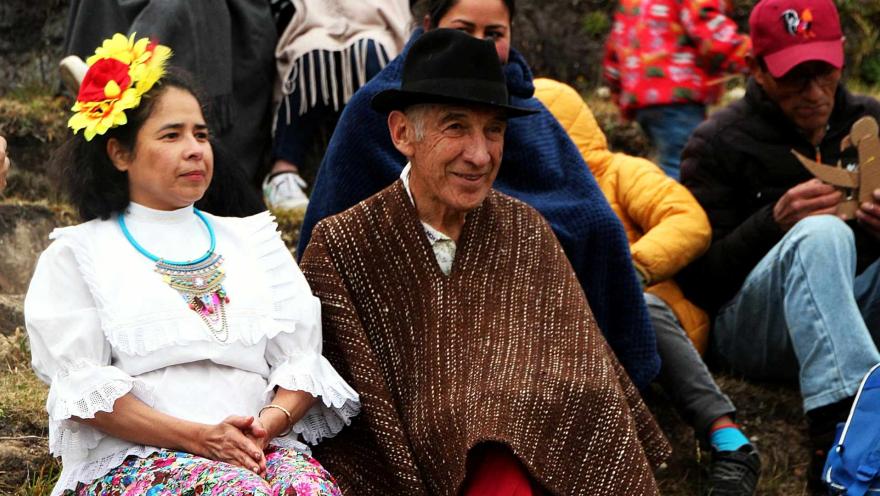 En el campo, un hombre, adulto mayor, y una mjujer campesina sentados sobre una montaña.