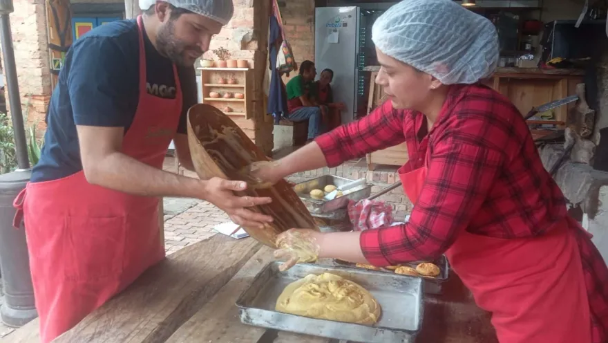  Un hombre y una mujer jóvenes cocinan con delantal y cofias unos amasijos. La cocina es artesanal.