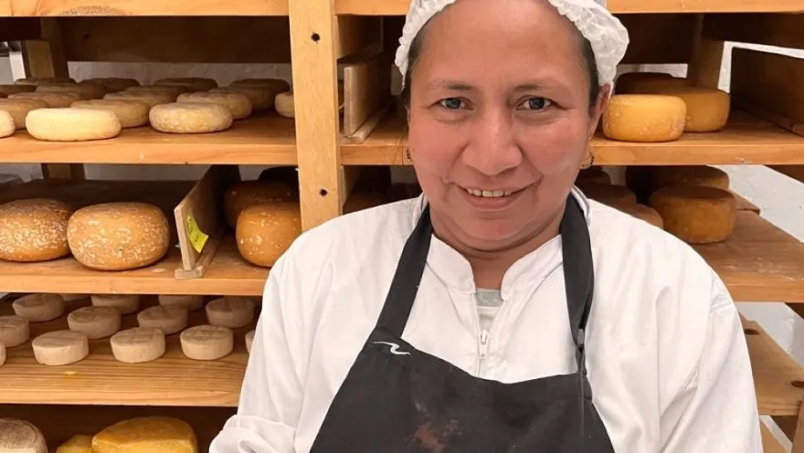 Mujer joven de tez canela sonriente con cofia delante de una estantería de madera con quesos.