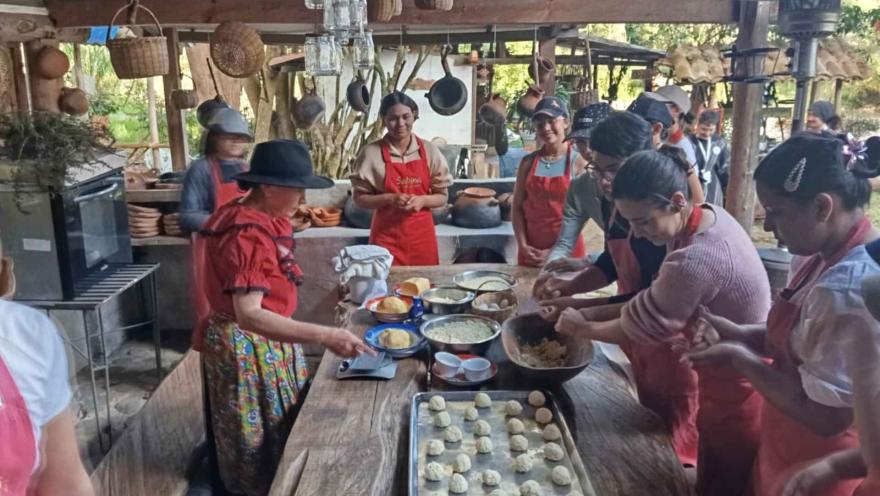  Mujer adulta con sombrero y vestido campesino enseña a cocinar a varias personas jóvenes en una concina artesanal. Hay una mesa con varios amasijos y harinas.