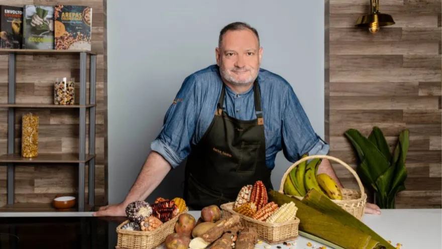 Chef Carlos Gavíria sonríe a la cámara, lleva camisa azul y delantal negro. Delante se observa una mesa con ingredientes como plátanos, papas, mazorcas y yucas. 