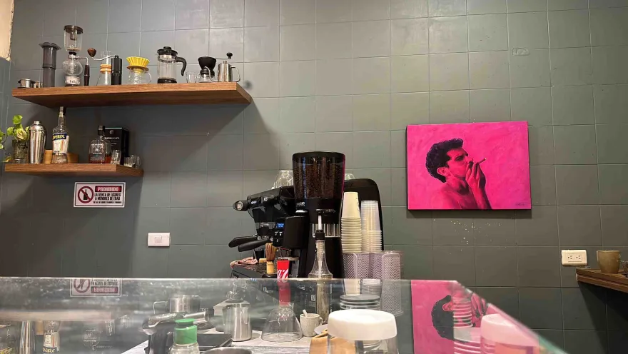 The interior of a kitchen with coffee-making equipment. In the background is a gray wall with a pink painting of a man smoking.