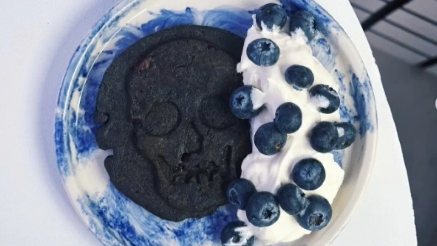 Postre con galleta, helado y arándanos. La galleta es negra y tieje forma de calabera. El postre está sobre un plato blanco con manchas azules.