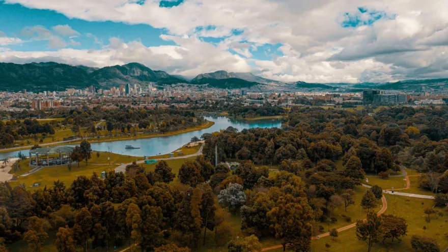 Simón Bolívar Park, a wide aerial view from a drone, showing the treetops, expansive green spaces, the enormous lake, city buildings in the background, and the eastern hills under a cloudy blue sky.