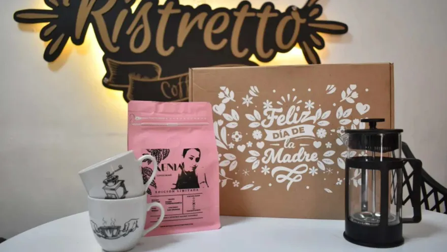 Una mesa blanca sobre la que estan dos pocillos, un paquete de café, una máquina para prepara café, una caja con un aviso que dice "feliz día de la madre" y al fondo sobre la pared el aviso que dice Ristretto.
