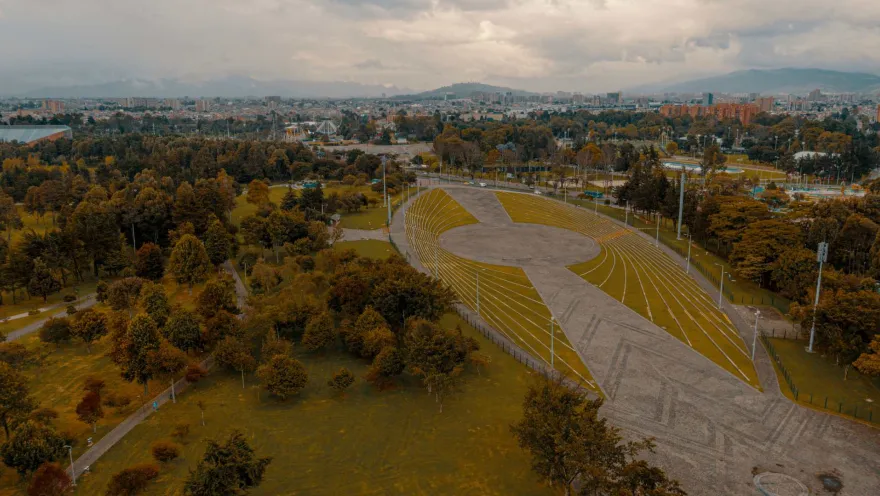 Simón Bolívar Park, a wide aerial view from a drone, showing the events plaza surrounded by leafy trees, with city buildings and a few mountains in the background.